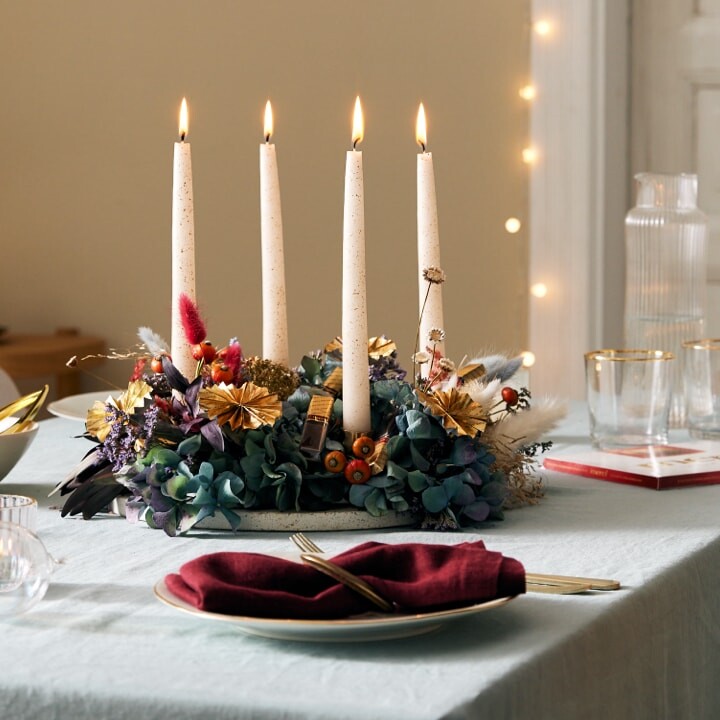 Auf einem Tisch ein Adventskranz aus Trockenblumen, merci Schokoladen-Stäbchen und vier langen weißen Kerzen