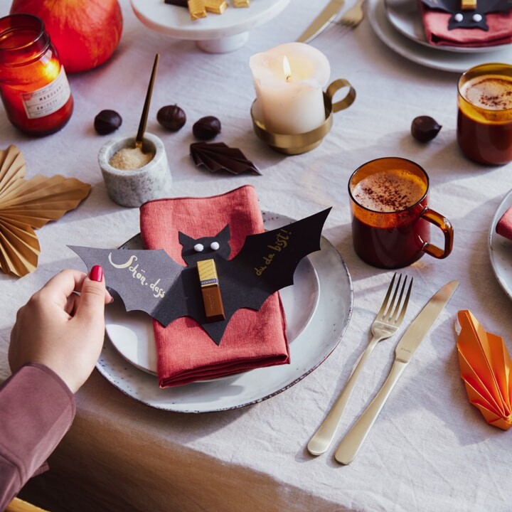 Tellerdeko. Aus schwarzer Pappe ausgeschnittene Fledermaus, mit aufgeklebten Augen, einem merci Schokoladen-Stäbchen und dem Schriftzug: Schön, dass du da bist!