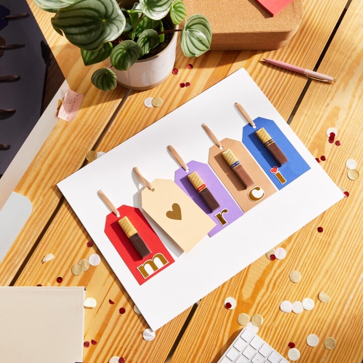 Carte artisanale sur laquelle sont disposées côte à côte cinq étiquettes cadeaux avec des bâtonnets de chocolat merci collés et le mot : merci